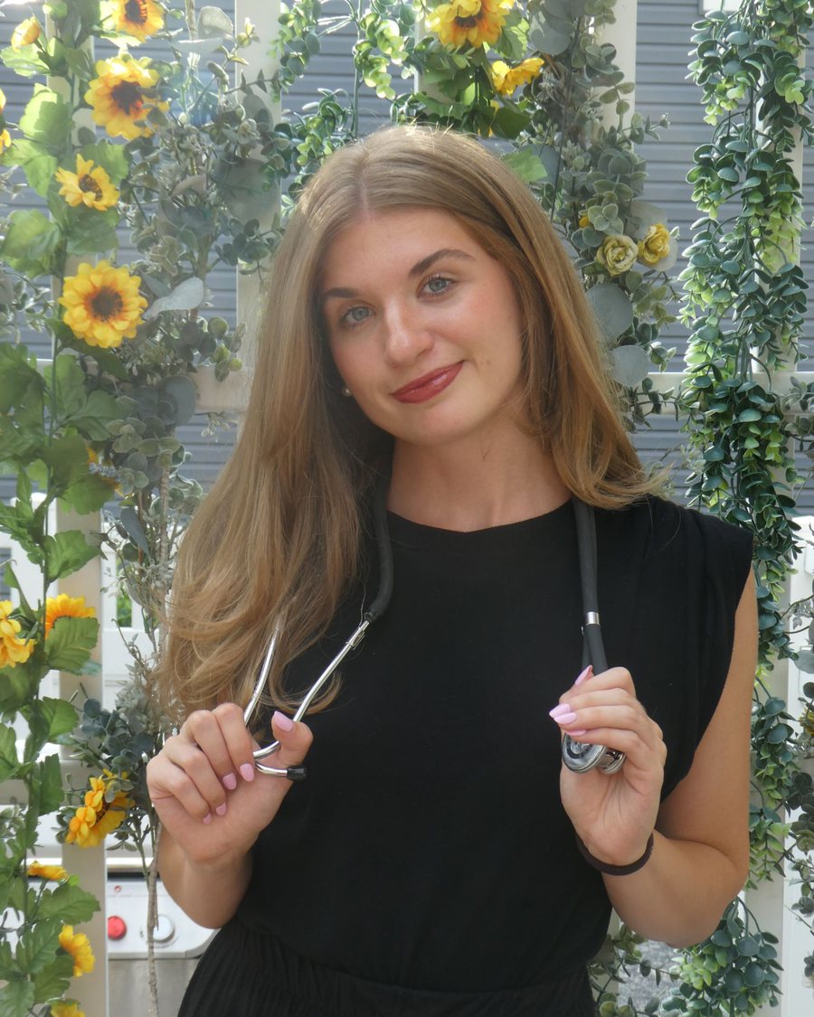 Elyse Fearon smiling, holding her stethoscope, in front of a wall of sunflowers and greenery
